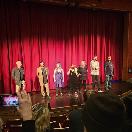 Sju personer står på en scen framför en röd ridå, med publik i förgrunden.