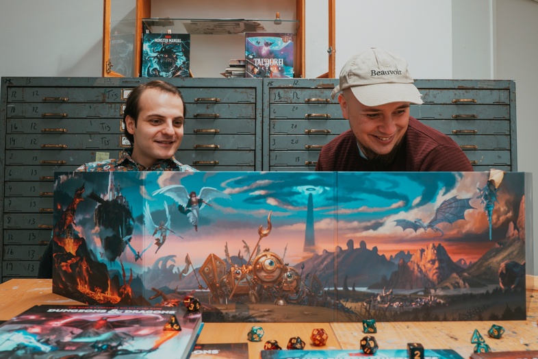 Två personer sitter vid ett bord med Dungeons & Dragons böcker, tärningar och spelledarskärm.