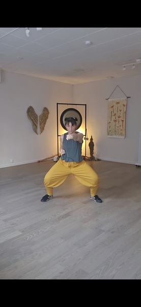 Person i gula byxor och grå topp gör en kampsportsposition i ett ljust rum med dekor och gong.