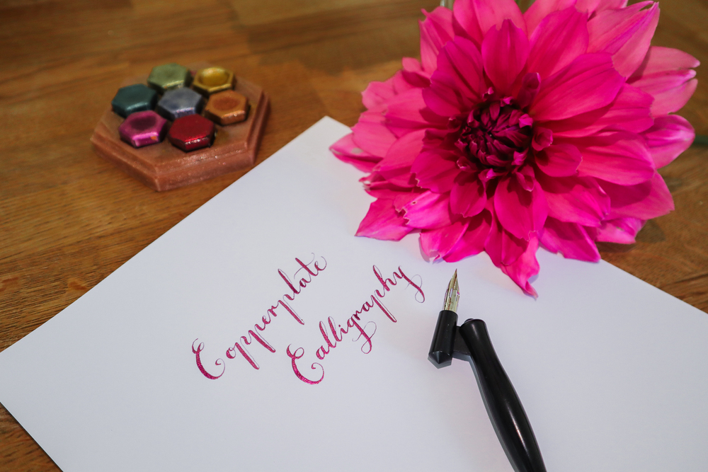 Ett papper med kalligrafitext, en bläckpenna, en stor rosa blomma och färgpalett på ett träbord.