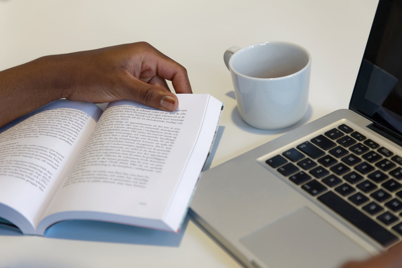 En hand bläddrar i en bok bredvid en bärbar dator och en vit kaffekopp på ett bord.