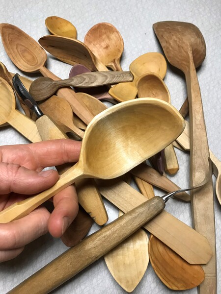 En hand håller en träslev ovanför en hög med olika handgjorda träskedar och snideriverktyg.