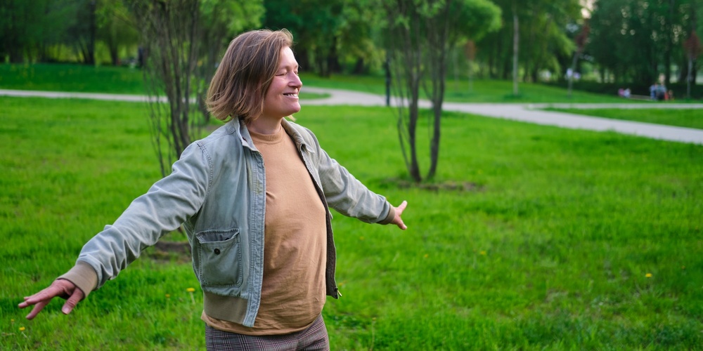 Person står med utsträckta armar i en grön park, ser glad ut och njuter av naturen.