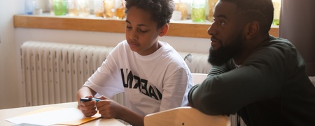 En pojke och en man sitter vid ett bord och arbetar tillsammans med papper och penna i ett ljust rum.