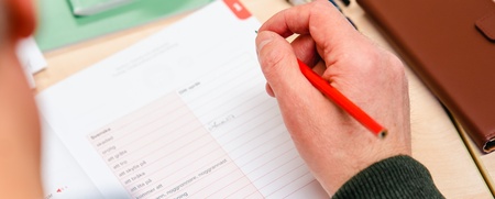 En person skriver med röd penna på ett papper vid ett skrivbord med anteckningsblock, pennor och fodral.