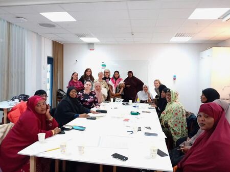 En grupp kvinnor sitter och står runt ett bord i ett ljust mötesrum, flera bär färgglada sjalar.