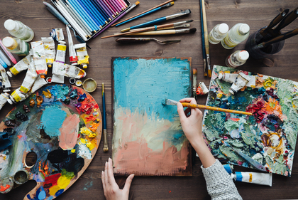 En person målar abstrakt konst på en duk med pensel, omgiven av färgtuber, penslar och färgpaletter på ett bord.