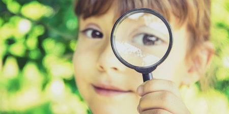 Ett barn håller upp ett förstoringsglas framför sitt öga utomhus, med grönskande bakgrund.
