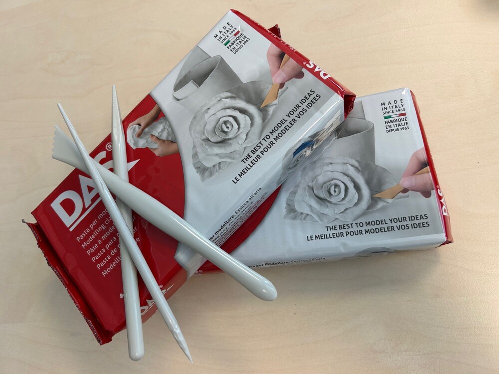 Två paket lufttorkande lera och tre vita modelleringsverktyg på ett ljust bord.