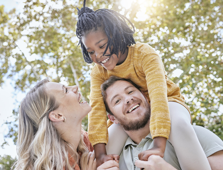 En glad familj utomhus, ett barn sitter på en vuxens axlar och alla ler mot varandra.