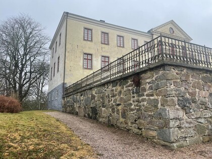 Stenmur och gammal gul byggnad med röda fönster, omgiven av träd och gräsmatta under en mulen himmel.