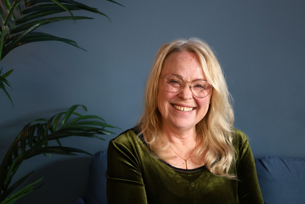 Leende äldre kvinna med långt blont hår och glasögon sitter mot blå vägg, grön tröja och växt i bakgrunden.