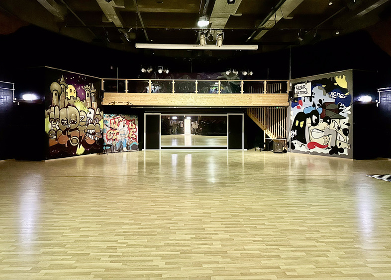 Stor danssal med trägolv, graffiti på väggarna och en balkong med räcke ovanför speglar.