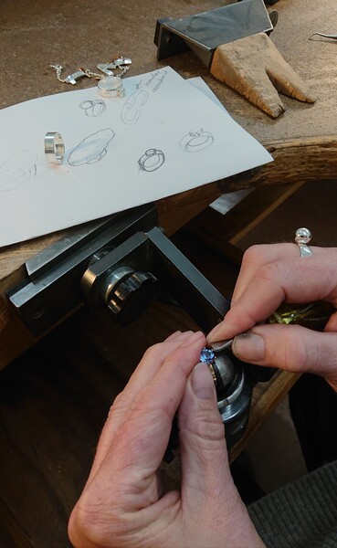 En person arbetar med att sätta en blå sten i en ring vid ett arbetsbord med smyckesritningar och verktyg.