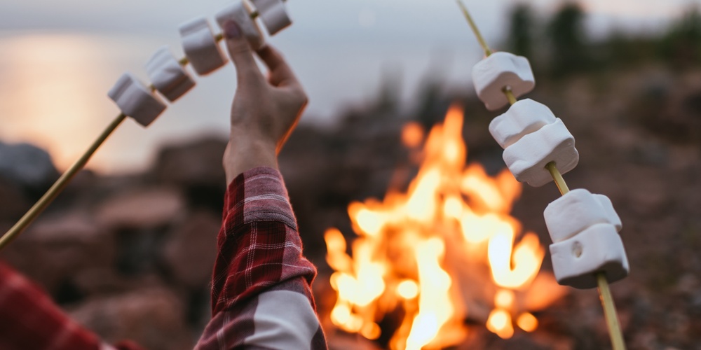 En person grillar marshmallows på pinnar över en öppen eld vid en sjö eller strand.