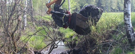 En grävmaskin gräver vid en liten bäck i en grönskande skogsmiljö.