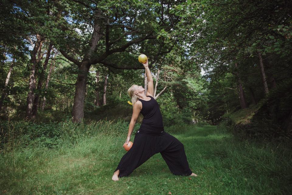 Person i svarta kläder gör yoga eller stretching med frukt i händerna i en grön skog.