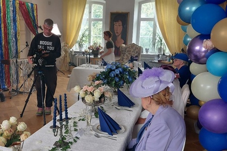 En bild av en filminspelning med en regissör, kameraman och två vackert klädda äldre damer som är skådespelare.