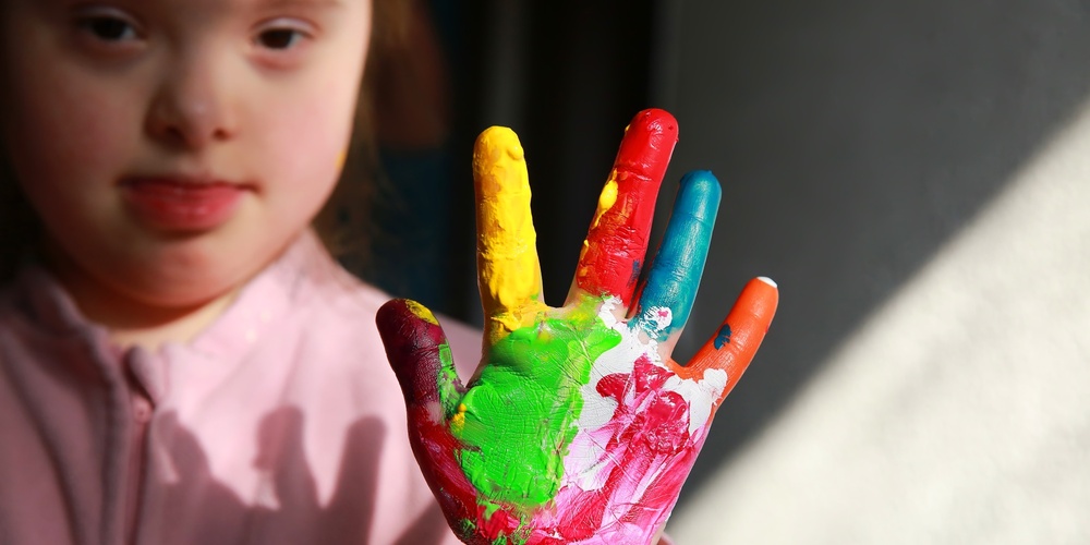 Ett barn visar sin hand täckt av färgglad fingerfärg, med ansiktet delvis synligt i bakgrunden.