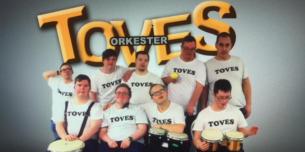 Grupp med åtta personer i vita TOVES-tröjor spelar trummor framför en stor TOVES-logga.