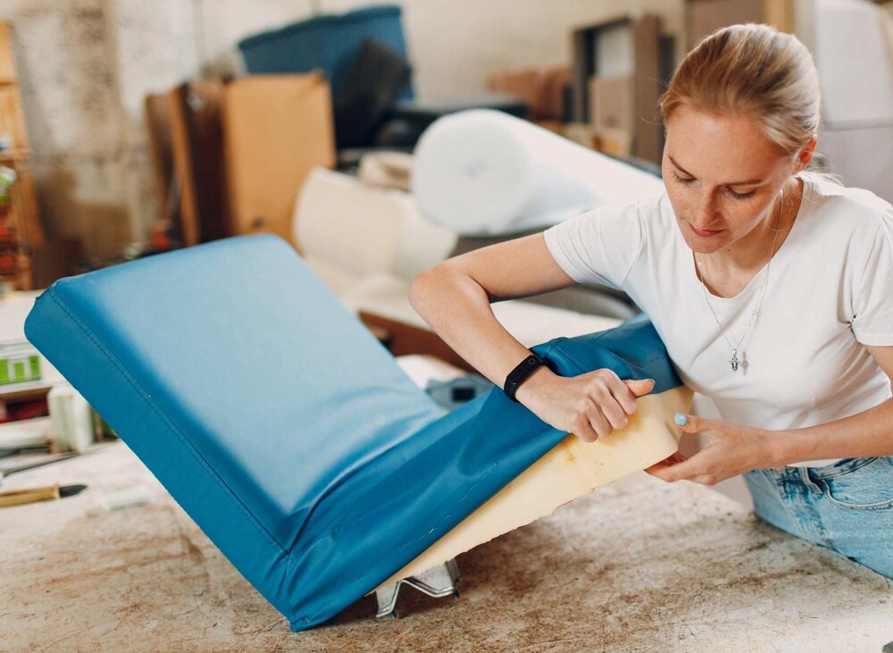 Kvinna klär om en blå stolsdyna med tyg i en verkstadsmiljö.