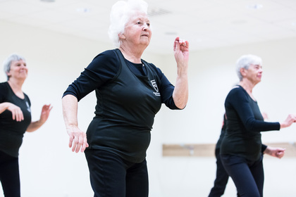 Äldre kvinnor deltar i en dans- eller träningsklass i ett ljust rum, klädda i svarta träningskläder.