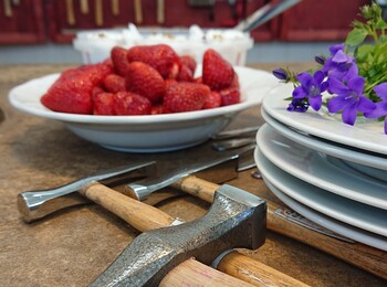 En närbild av hammare, tallrikar, jordgubbar och blommor på ett bord i en verkstadsmiljö.