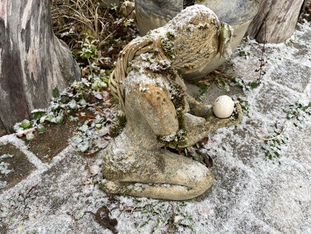 En mossig staty av en knästående figur med långt hår håller en rund sten, täckt av lätt snö utomhus.
