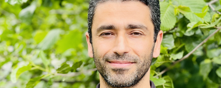Man med mörkt hår och skägg står utomhus framför gröna träd och bär en mörk pikétröja.