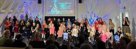 En grupp barn och vuxna står på en scen inför publik, med färgglad ljussättning och juldekorationer.