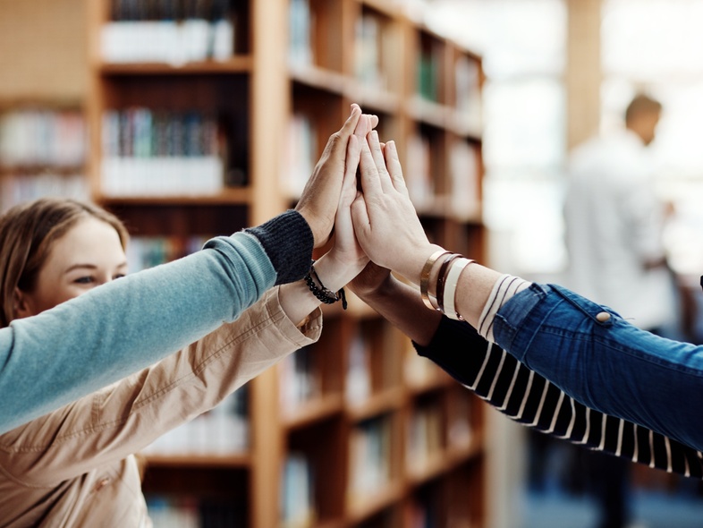 Fyra unga personer gör en gemensam high five i ett bibliotek, glada och engagerade.