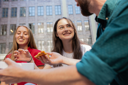 Tre unga personer sitter utomhus och äter pizza tillsammans, glada och avslappnade.