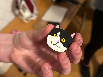 En hand håller en liten figur av en svartvit katt med gula ögon, i ett rum med trägolv och strykjärn i bakgrunden.