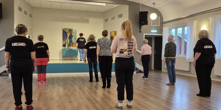 En grupp personer står i ett dansrum och följer en instruktör på en liten scen längst fram.