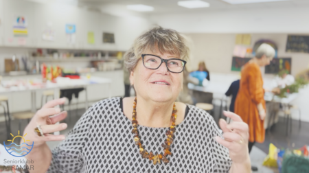 Äldre kvinna med glasögon och halsband gestikulerar i ett ljust rum med andra personer i bakgrunden.