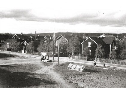 Svartvit bild av en liten by med trähus, grusvägar och några människor som promenerar.