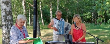 Tre personer spelar musik utomhus bland träd, en med keyboard, en med dragspel och en med papper.