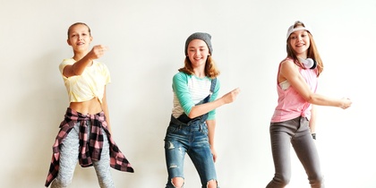 Tre ungdomar dansar energiskt i ett ljust rum med trägolv och vit vägg.