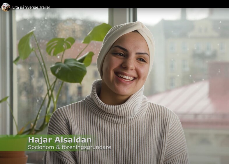 Leende kvinna i ljus hijab och stickad tröja sitter vid ett fönster med växter i bakgrunden.