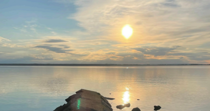 Solnedgång över en lugn sjö med en stenig brygga i förgrunden och mjuka moln på himlen.