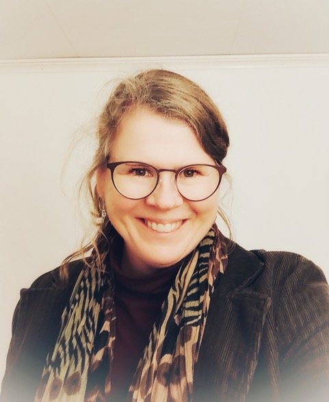 Leende kvinna med glasögon, brun kavaj och mönstrad halsduk mot ljus bakgrund.
