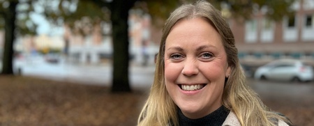 Leende kvinna står utomhus på en höstig gata med träd och löv i bakgrunden.