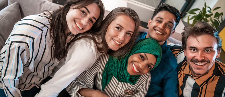 Fem unga vuxna sitter nära varandra och ler mot kameran i en vardagsrumsmiljö.