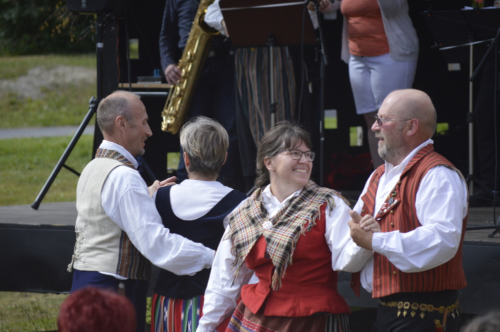 Fyra personer i traditionella folkdräkter dansar utomhus framför en scen med musiker.