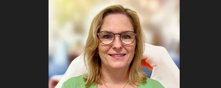 Kvinna med blont hår och glasögon ler mot kameran, bär grön tröja, suddig bakgrund.