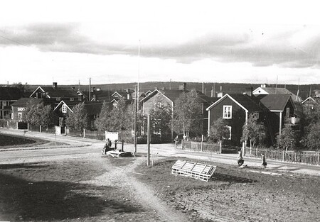 Svartvit bild av en liten by med trähus, grusvägar och några människor som promenerar.