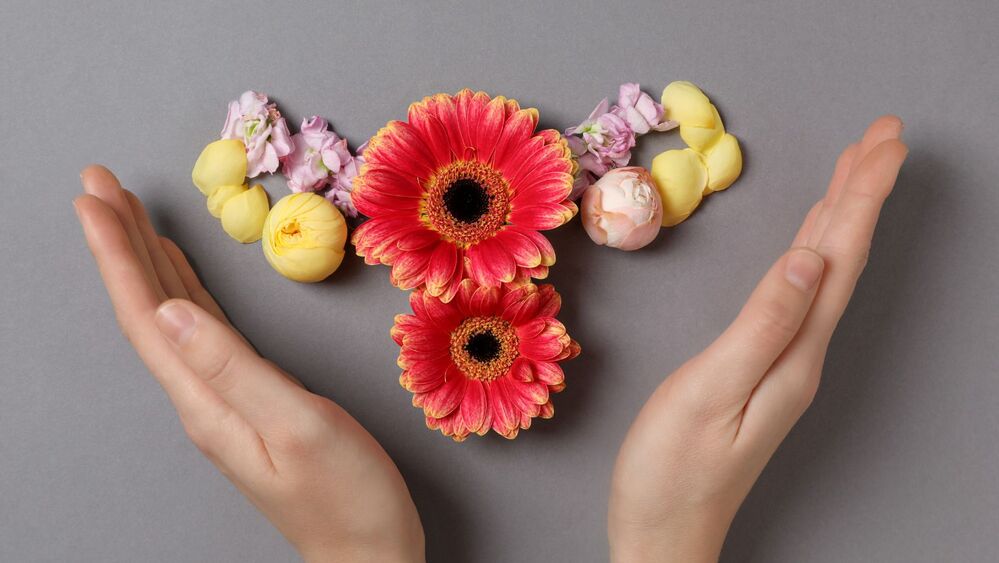 Två händer omger blommor arrangerade som en livmoder på grå bakgrund.