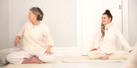 Två personer sitter på yogamattor i ett ljust rum och gör en sittande vridning.