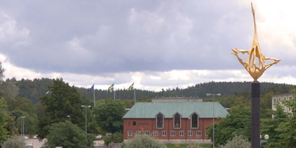 Stadsvy med en guldfärgad skulptur i förgrunden, grönska och en tegelbyggnad i bakgrunden.