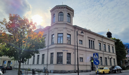 Gammal ljusgrå byggnad med torn, träd och bilar i förgrunden, sol bakom moln.